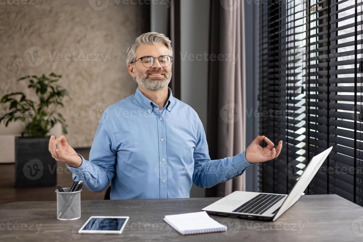 Confident mature man meditating at his office desk with a laptop and tablet, promoting mindfulness and relaxation in a work environment. photo