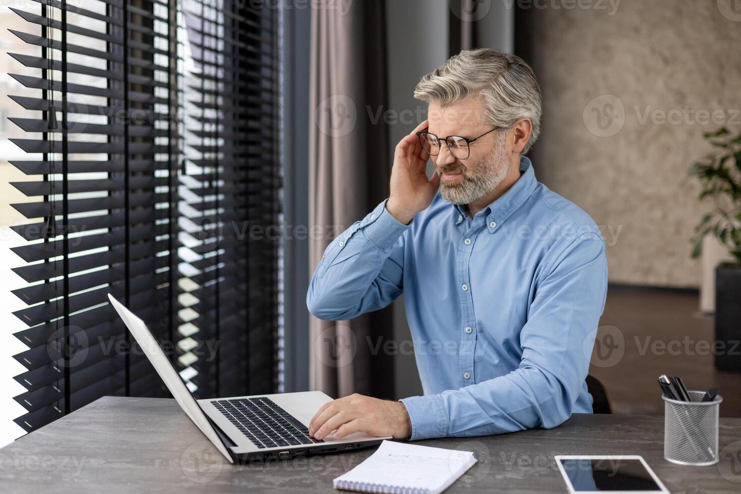 Middle-aged man with glasses and beard experiencing head pain while working on laptop at office desk. Concept of work stress and headache. photo