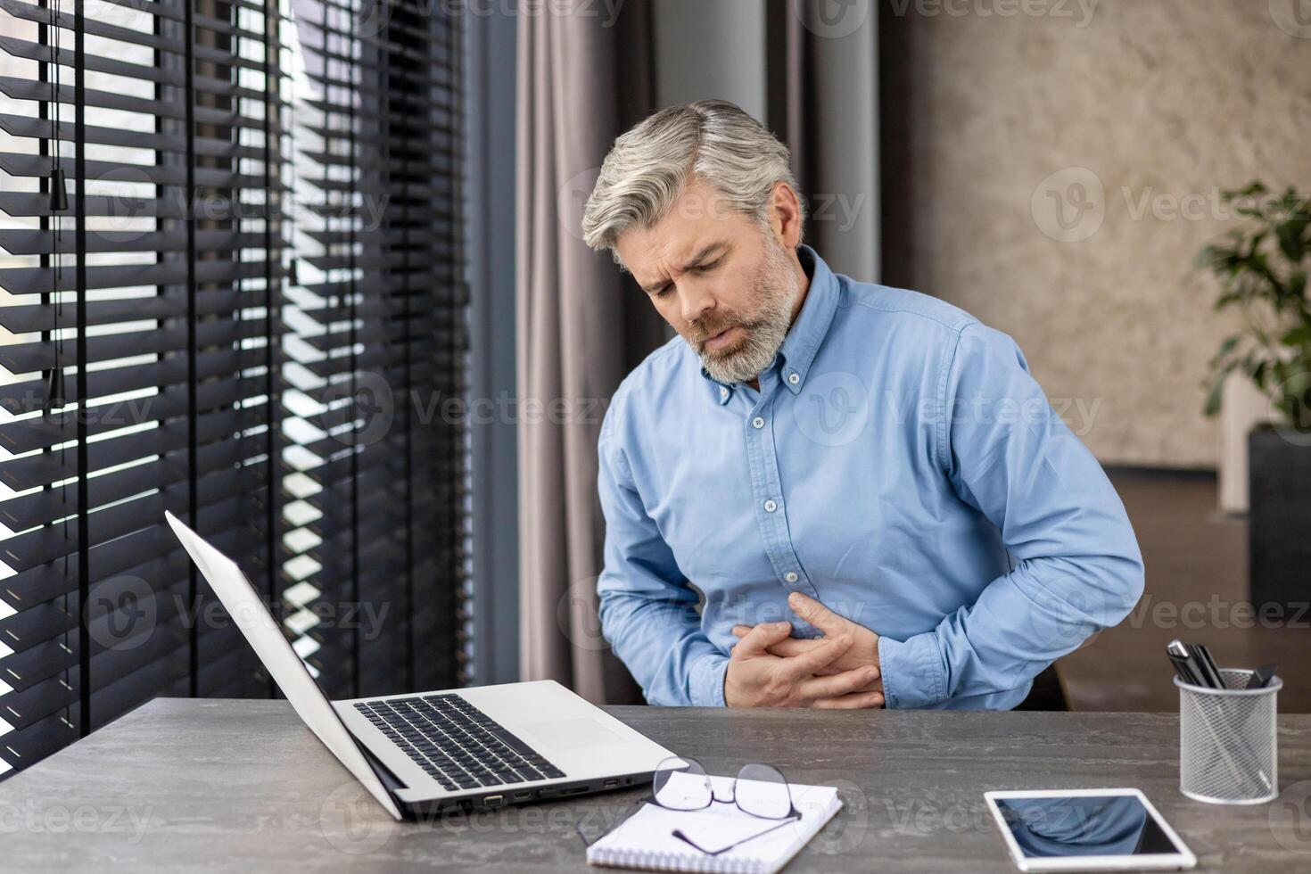 Businessman experiencing stomach pain while working on a laptop at a modern office desk. Stress, health issues, and discomfort. photo
