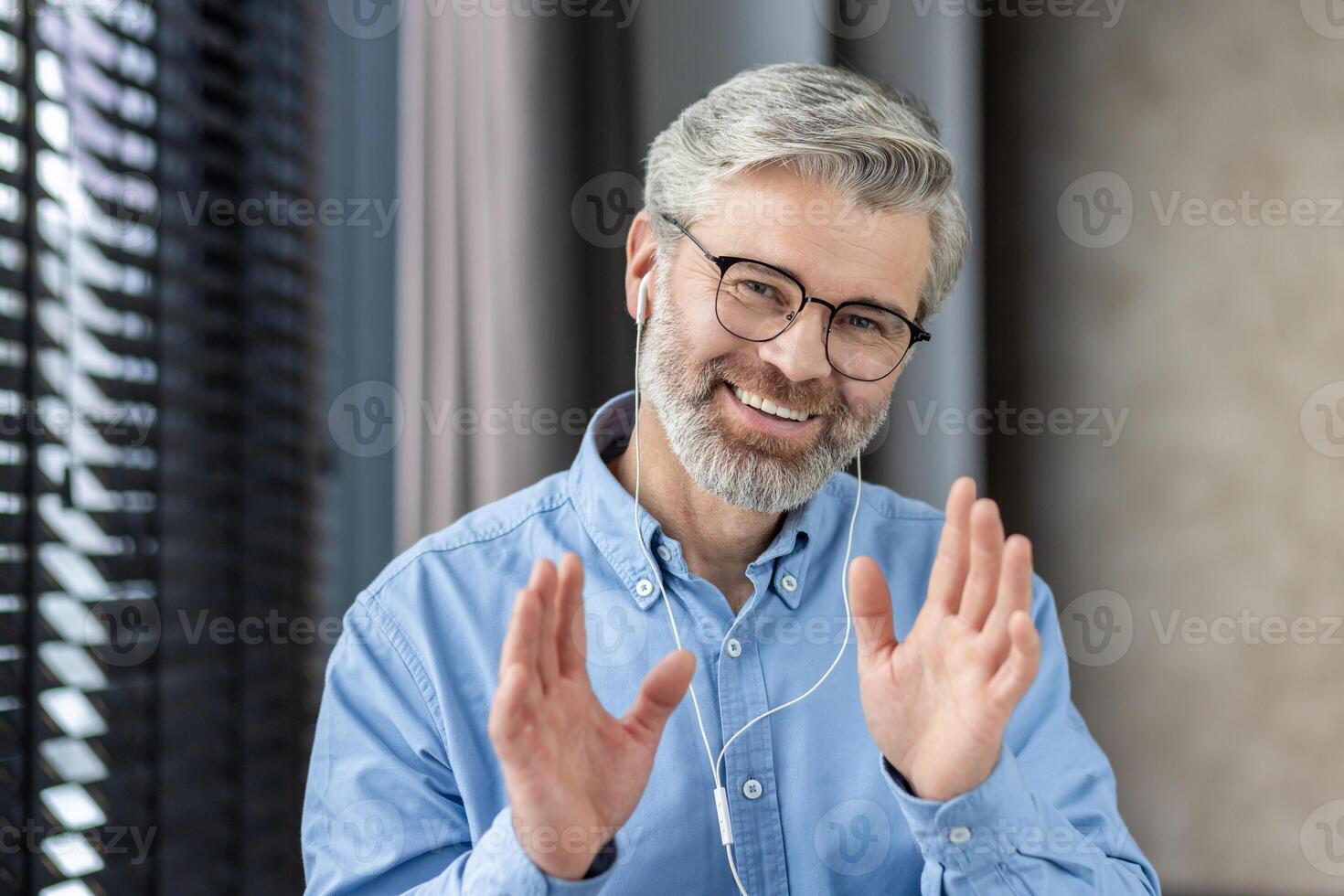 Senior man in an office setting wearing headphones, smiling and waving during a call. The man appears to be calm and happy as he engages in a remote work meeting. photo