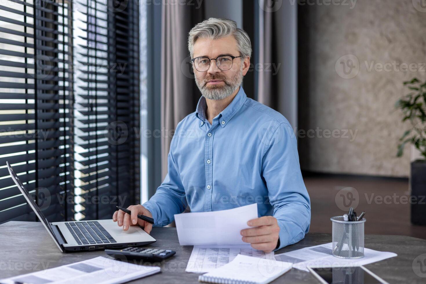 Middle-aged businessman wearing glasses and a blue shirt, analyzing documents at his desk in a modern office. He appears confident and professional, with a laptop, calculator, and paperwork photo