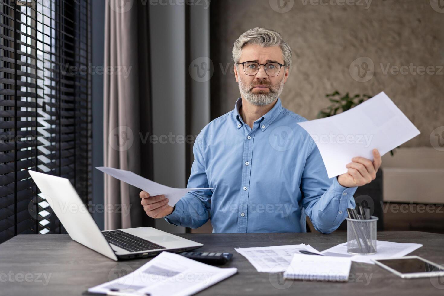 Professional businessman sitting at desk holding documents, displaying concern while working on laptop in modern office. Concept of work stress, business finance, office environment photo