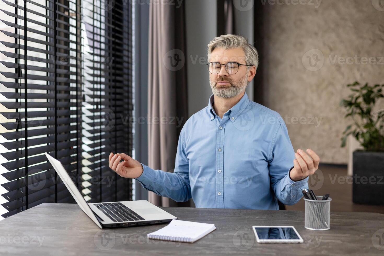 Businessman practicing meditation at his desk in a modern office setting with a laptop and tablet, promoting relaxation and mindfulness. photo