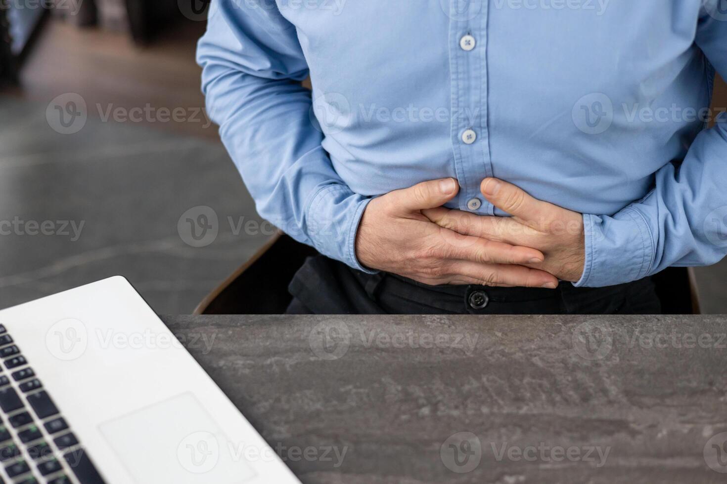 Businessman in blue shirt experiencing stomach pain while working on laptop at office desk, highlighting health issues related to stress or digestion. photo