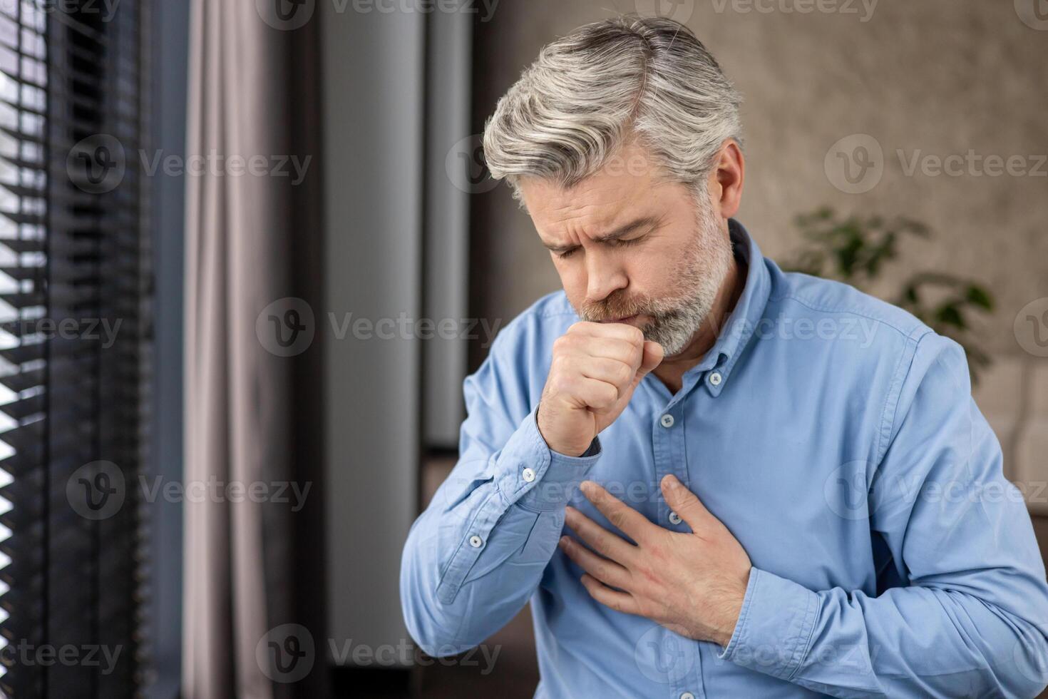Elderly man with grey hair and beard coughing and holding chest with painful expression. Health concept of illness, bronchitis, lung disease, or flu. Indoors with window and blurred background. photo