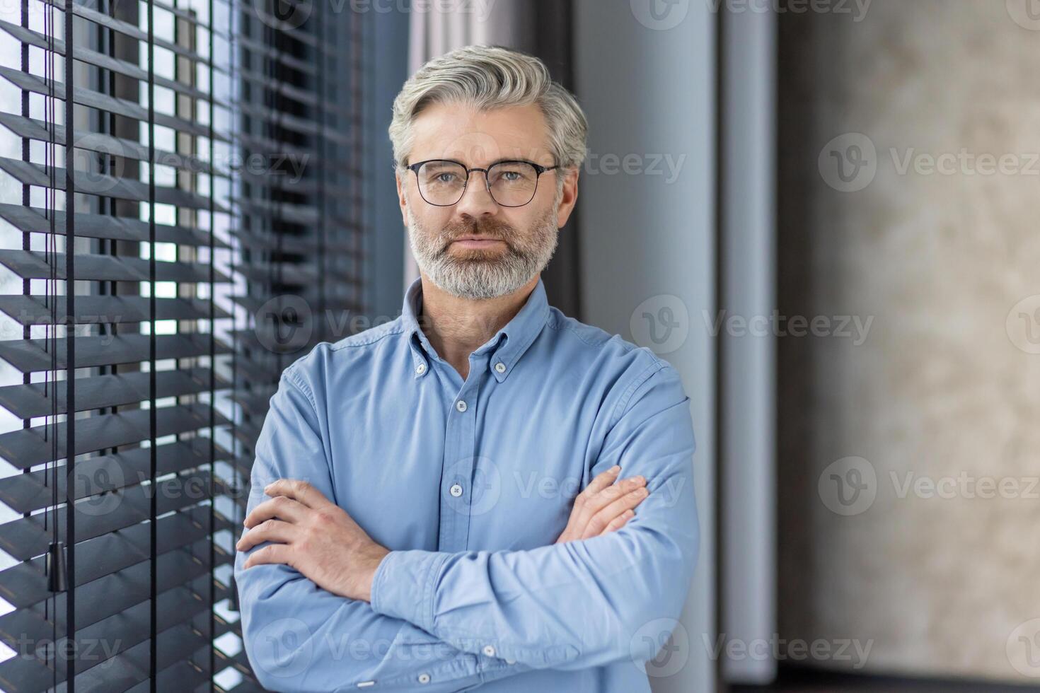 Portrait of caucasian businessman in eyeglasses standing with folded hands on apartment background. Successful aged male feeling calm and confident about future of company and economical stability. photo