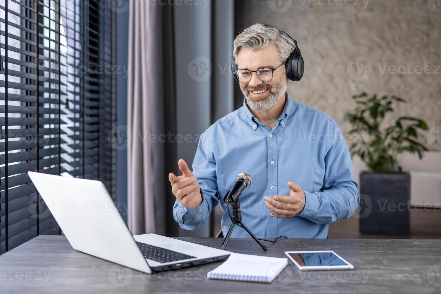 A gray-haired senior man is sitting at the table at home, wearing headphones in front of a microphone and chatting online via call, recording a webinar. photo