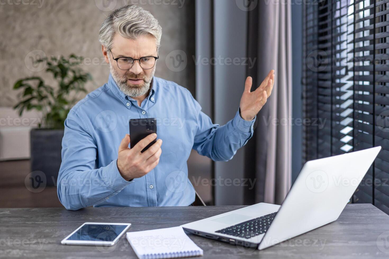 Sad disappointed boss businessman at workplace received online message email on phone, senior mature gray haired man using app on smartphone, inside office with laptop. photo