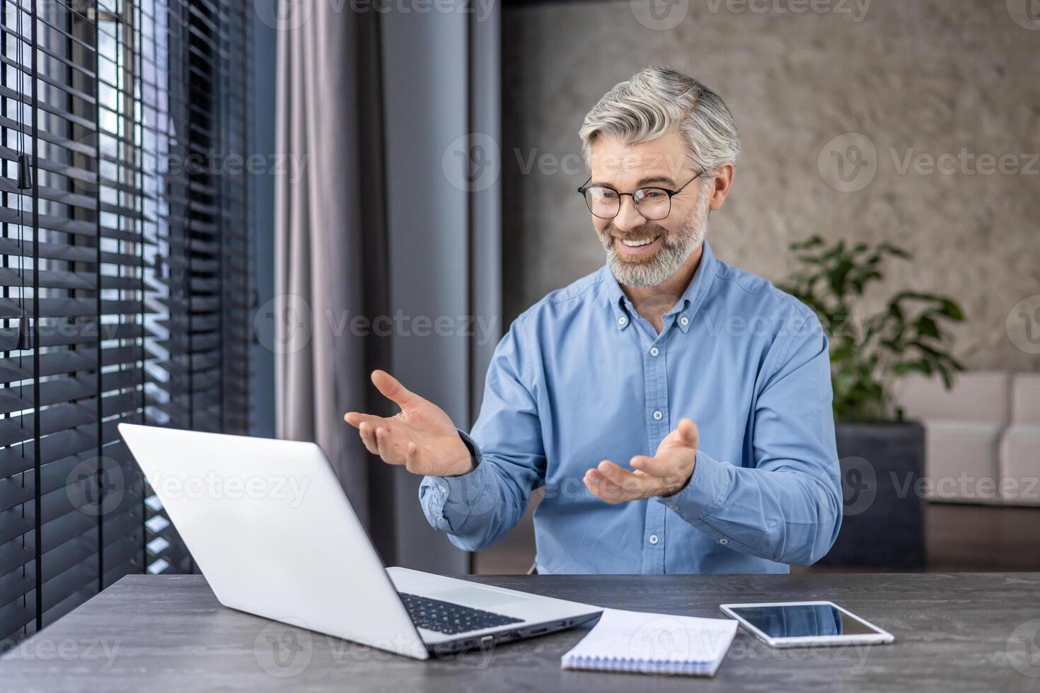 Smiling mature man with glasses in a blue shirt having a call on a laptop in a modern office setting, expressing positivity. photo