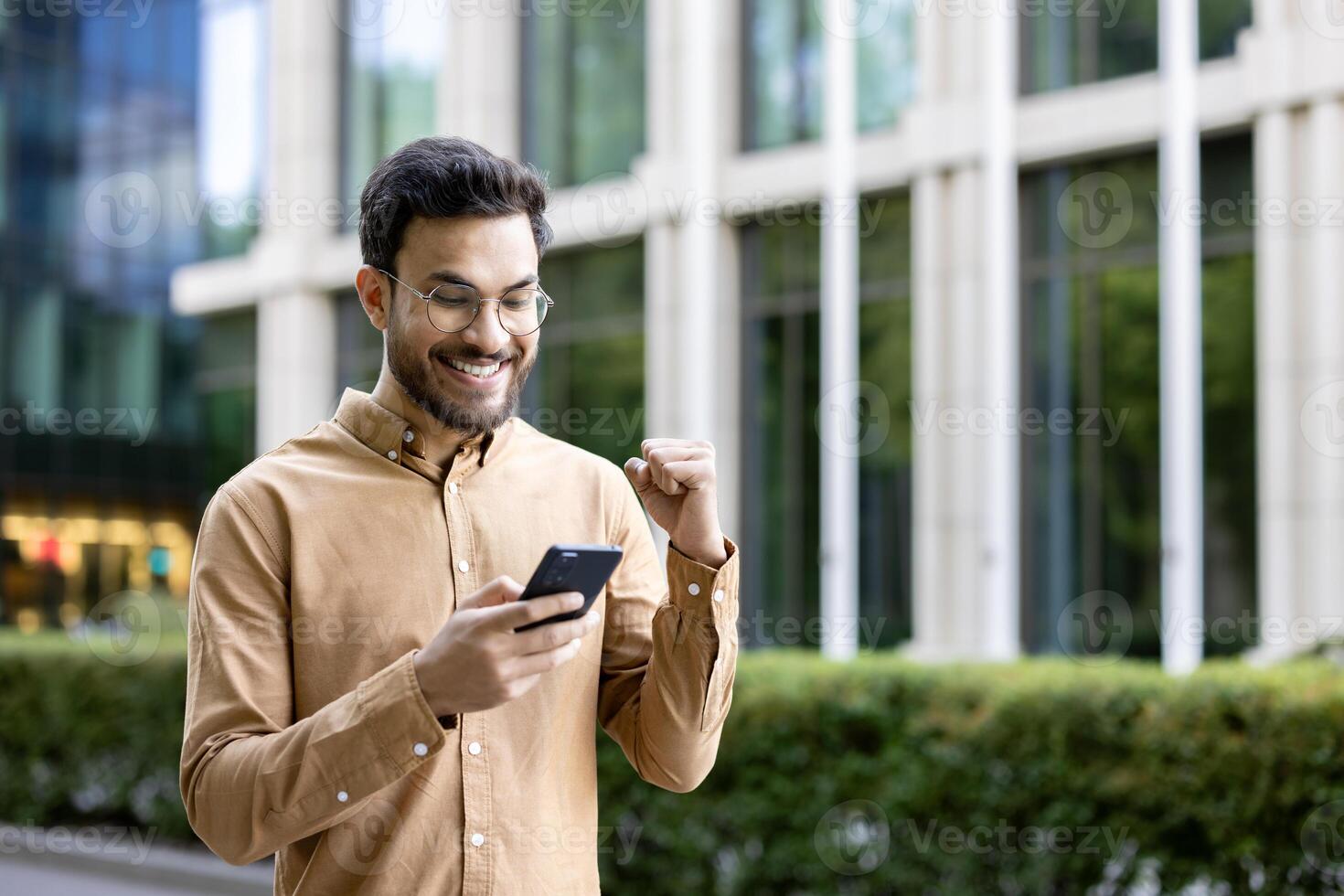 Young man in glasses smiling and celebrating success while using smartphone outdoors. Business professional receiving good news, feeling happy. Modern office building background. photo