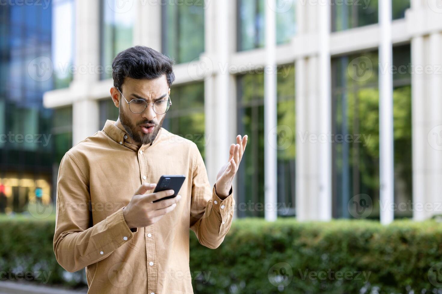 Man in casual clothing outside office building looking at smartphone with frustration. Business professional experiencing issue or news causing surprise or anger. photo