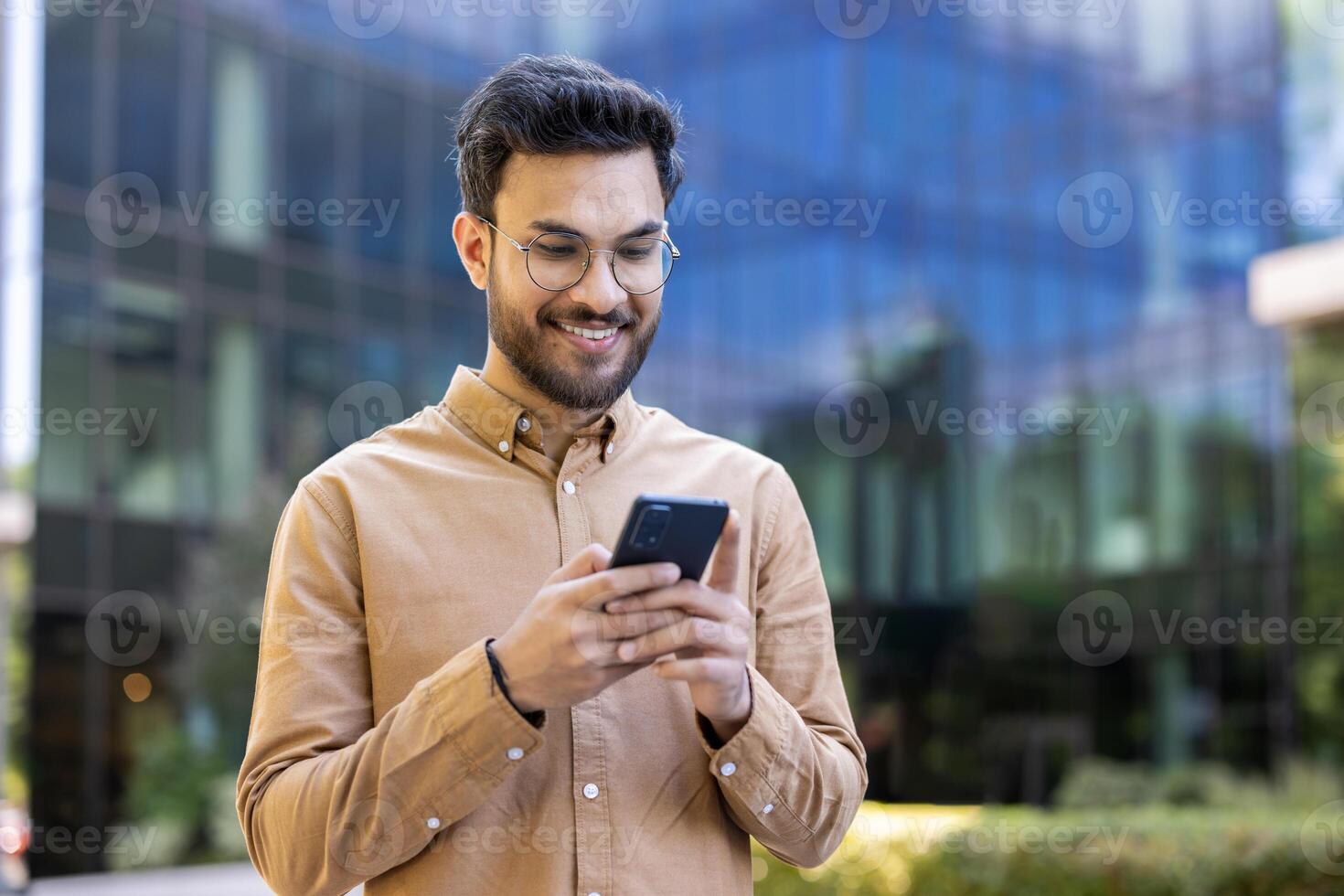 Smiling young man holding smartphone outdoors in urban setting. Represents connection, technology, communication, displaying positive feeling while interacting with phone near modern office building. photo