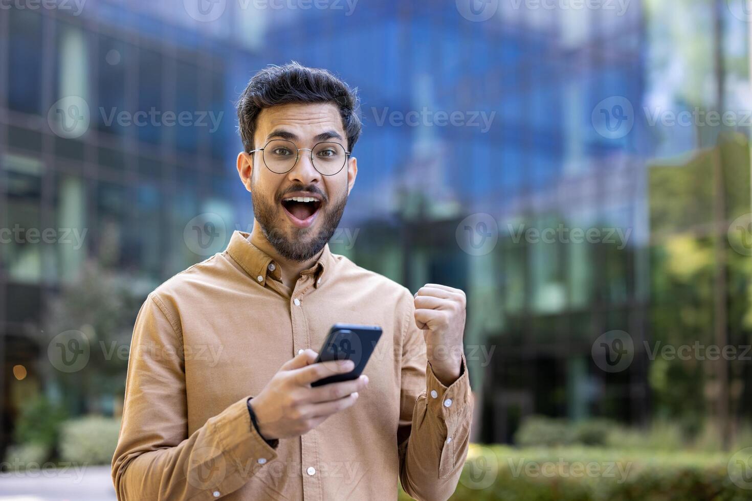 Businessman outdoors celebrating achievement with phone. Young professional looking at camera with excitement, expressing surprise and joy. Success moment captured with modern technology photo
