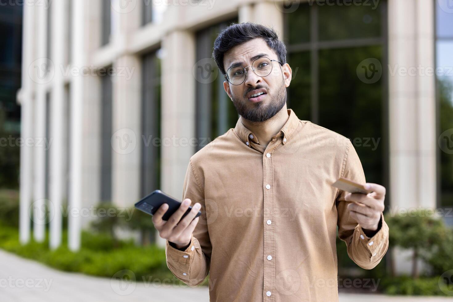 Man with glasses standing outside, holding phone and credit card, showing confused expression. Concept of financial uncertainty, technology frustration, decision making, or communication issues. photo