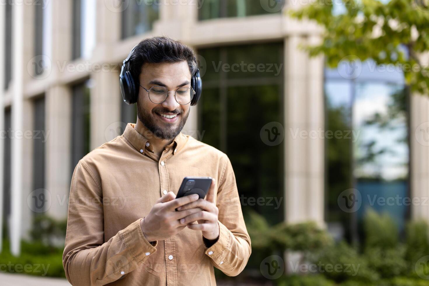 Confident man wearing headphones looking at smartphone outside modern urban building. He is relaxed and happy, enjoying music or engaging with friends or colleagues. photo