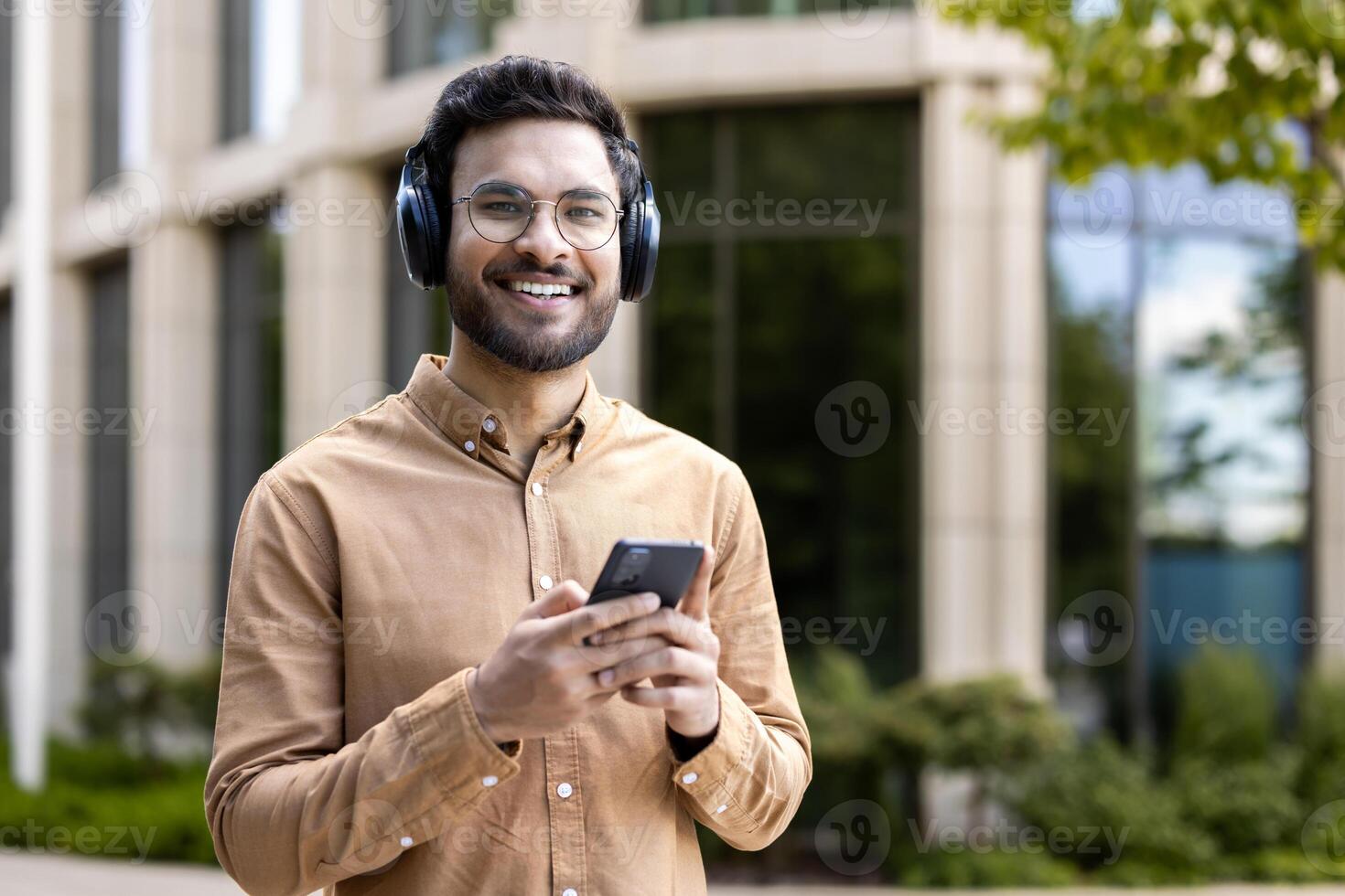 Smiling young man listens to music on headphones while holding smartphone in urban setting. Enjoys leisure time outdoors, expressing happiness and confidence. photo