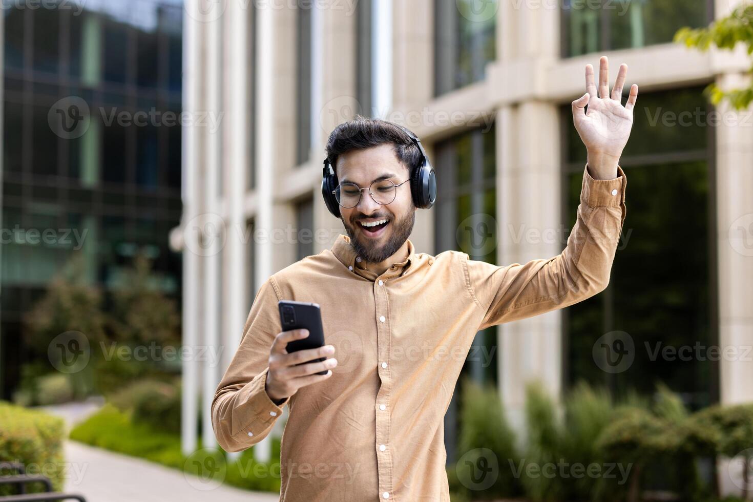 Smiling man wearing headphones uses smartphone enjoying music outdoors. Standing with hand raised in excitement, expressing happiness and enjoyment in sunny urban setting. photo