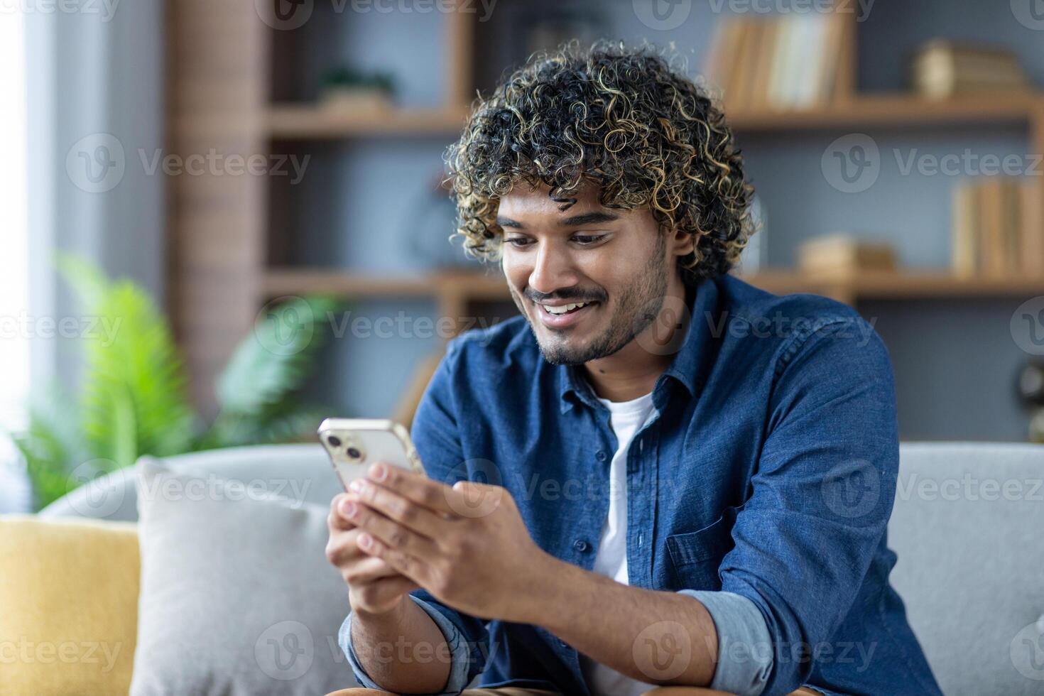 Young joyful man uses phone while sitting at home on sofa in living room of apartment, smiles happily, browses social media, and types text message. photo