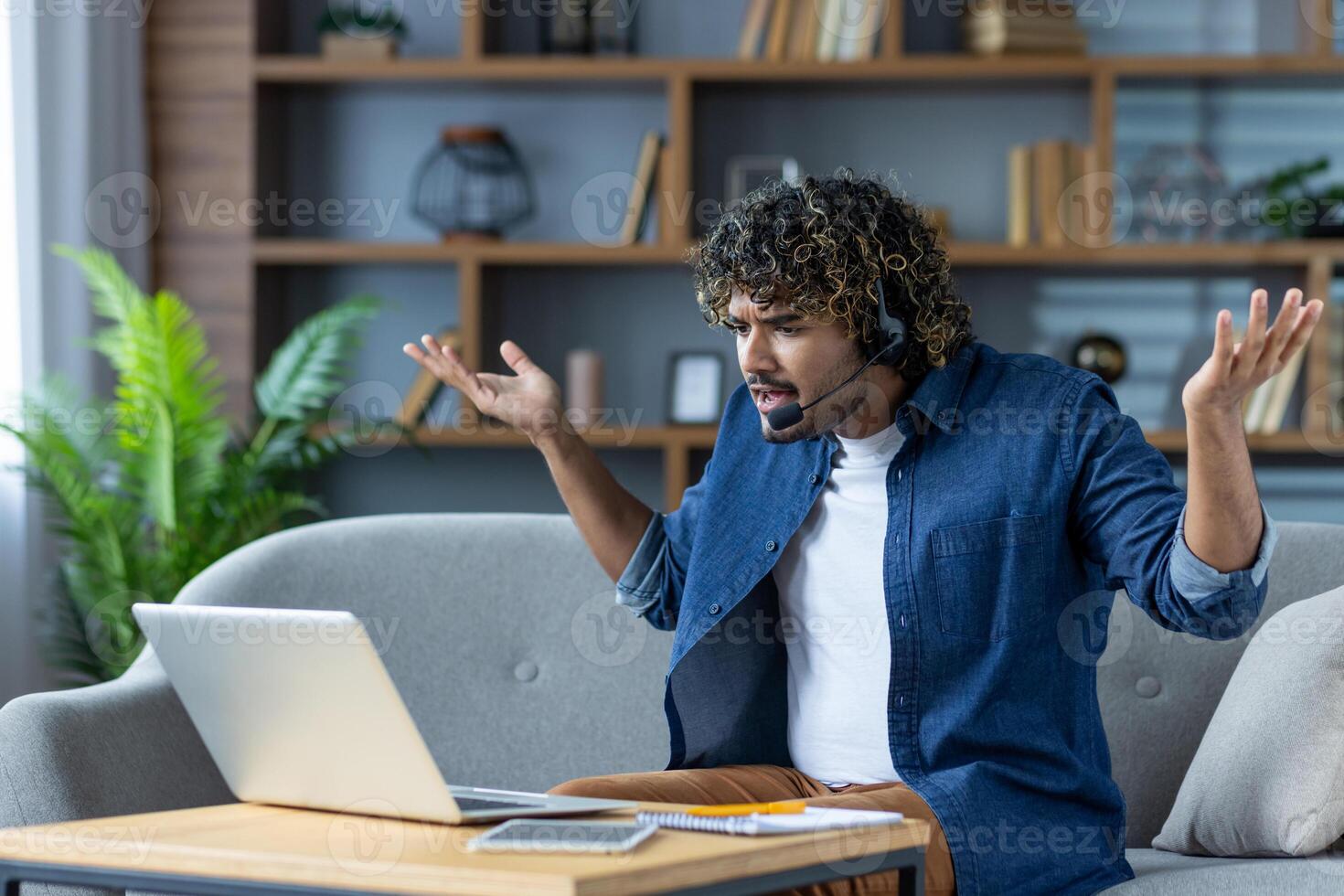 Dissatisfied man sitting on sofa at home, with headset and headphones. Nervous freelancer working remotely using call, arguing and shouting. photo