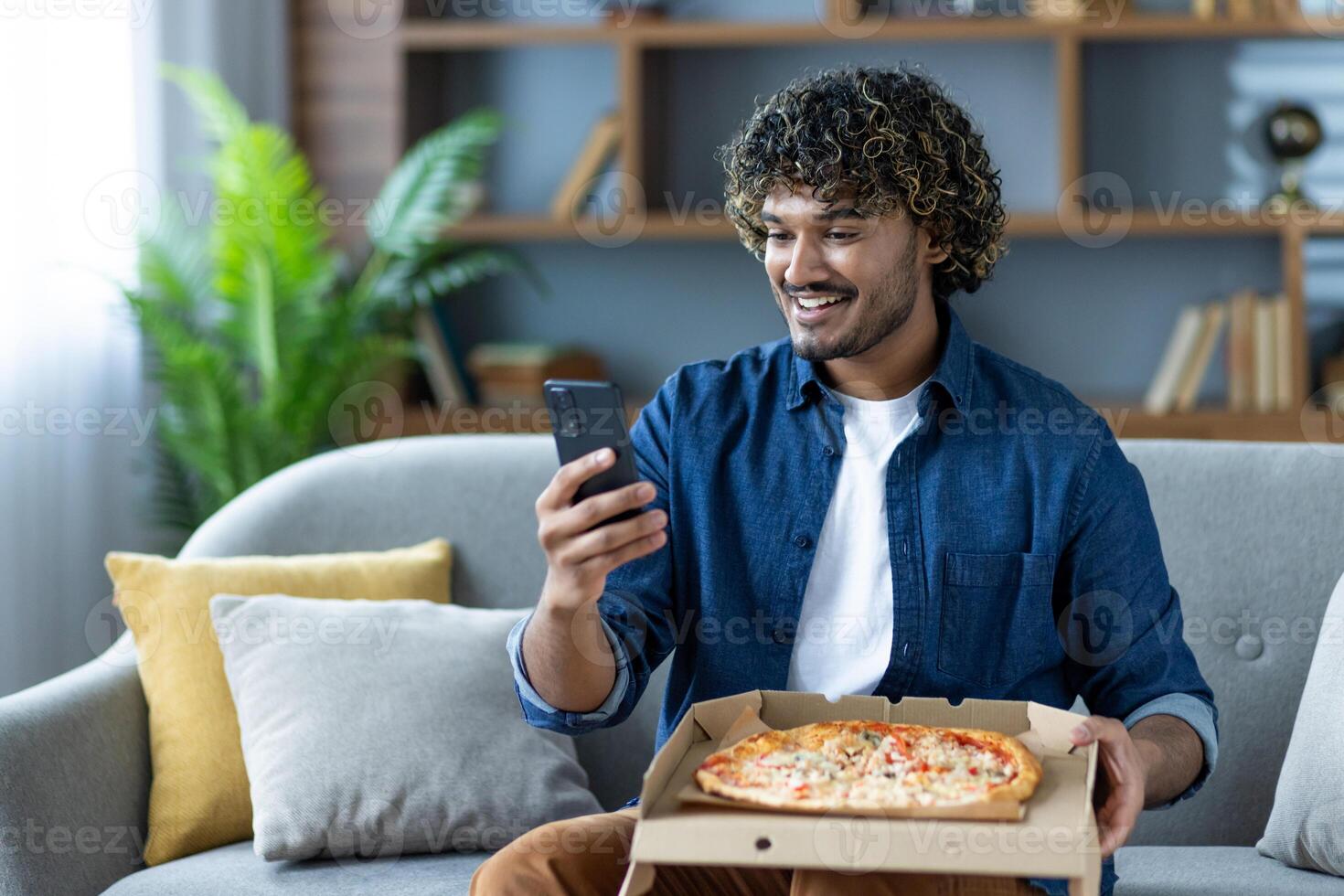 Young man sitting at home with pizza and phone in hands, satisfied with quality of online order, and speed of delivery. Happy customer ordered on sofa in living room. photo