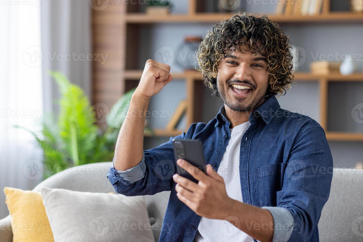 Portrait of joyful successful man with phone in hands. Winner looking at camera celebrating triumphant results of achievement, sitting on sofa in living room. photo