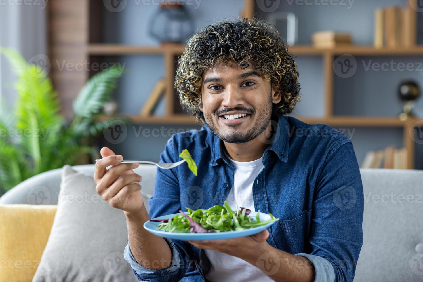 A cheerful man sits comfortably on a sofa, enjoying a fresh green salad. His expression of happiness highlights a healthy lifestyle. The cozy living room setting adds warmth and relaxation. photo