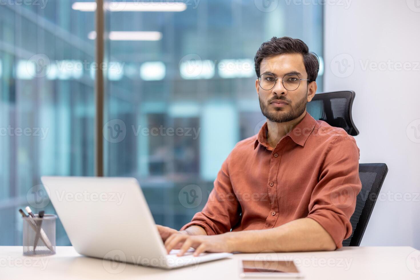 Confident businessman working at laptop in modern office. Man in casual attire focused on tasks. Bright, professional workspace with large windows. Concept of technology, business, and productivity. photo