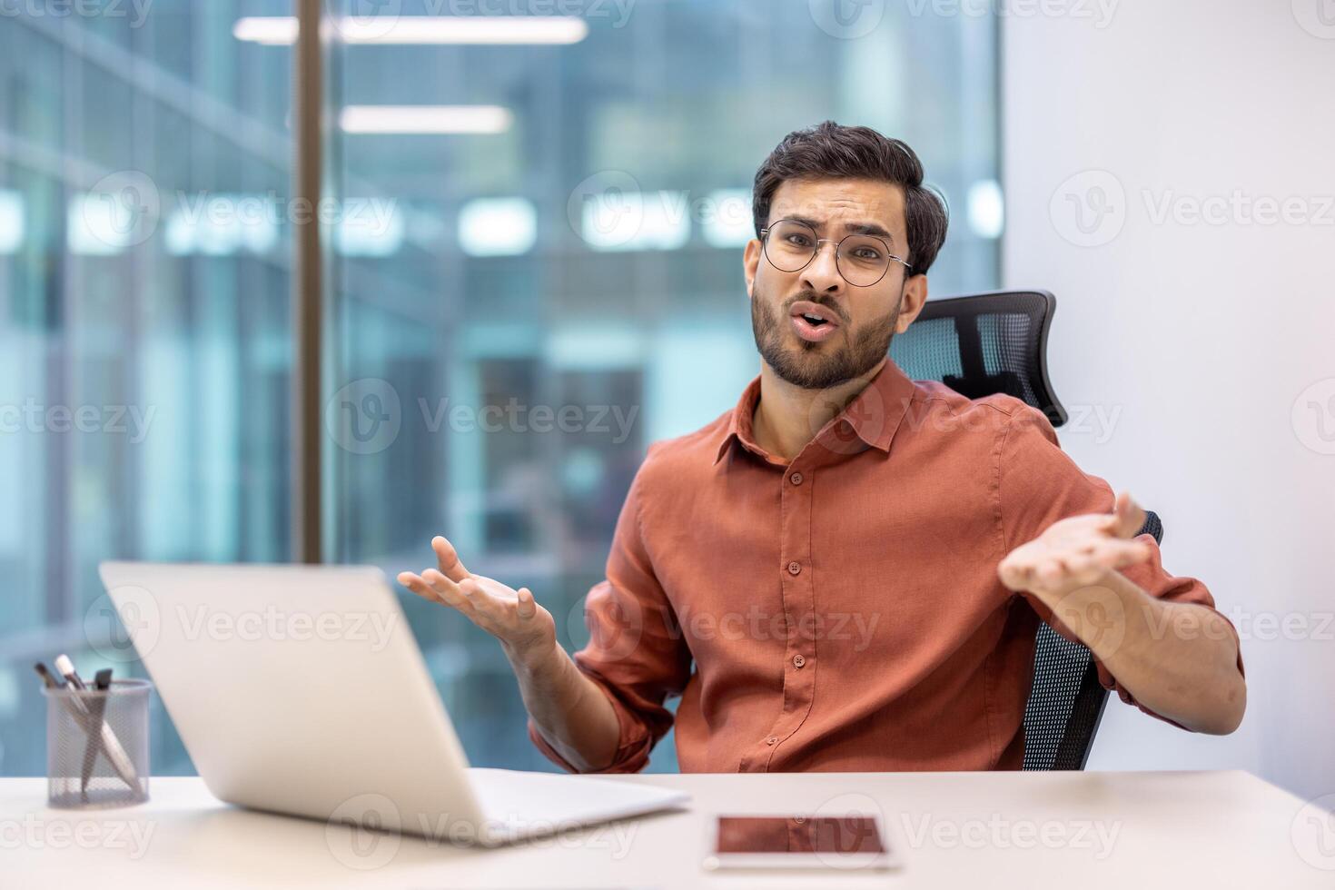 Confused office worker gesturing at desk with laptop and smartphone. Man in office frustrated, expressing emotions at work during business meeting or call. photo