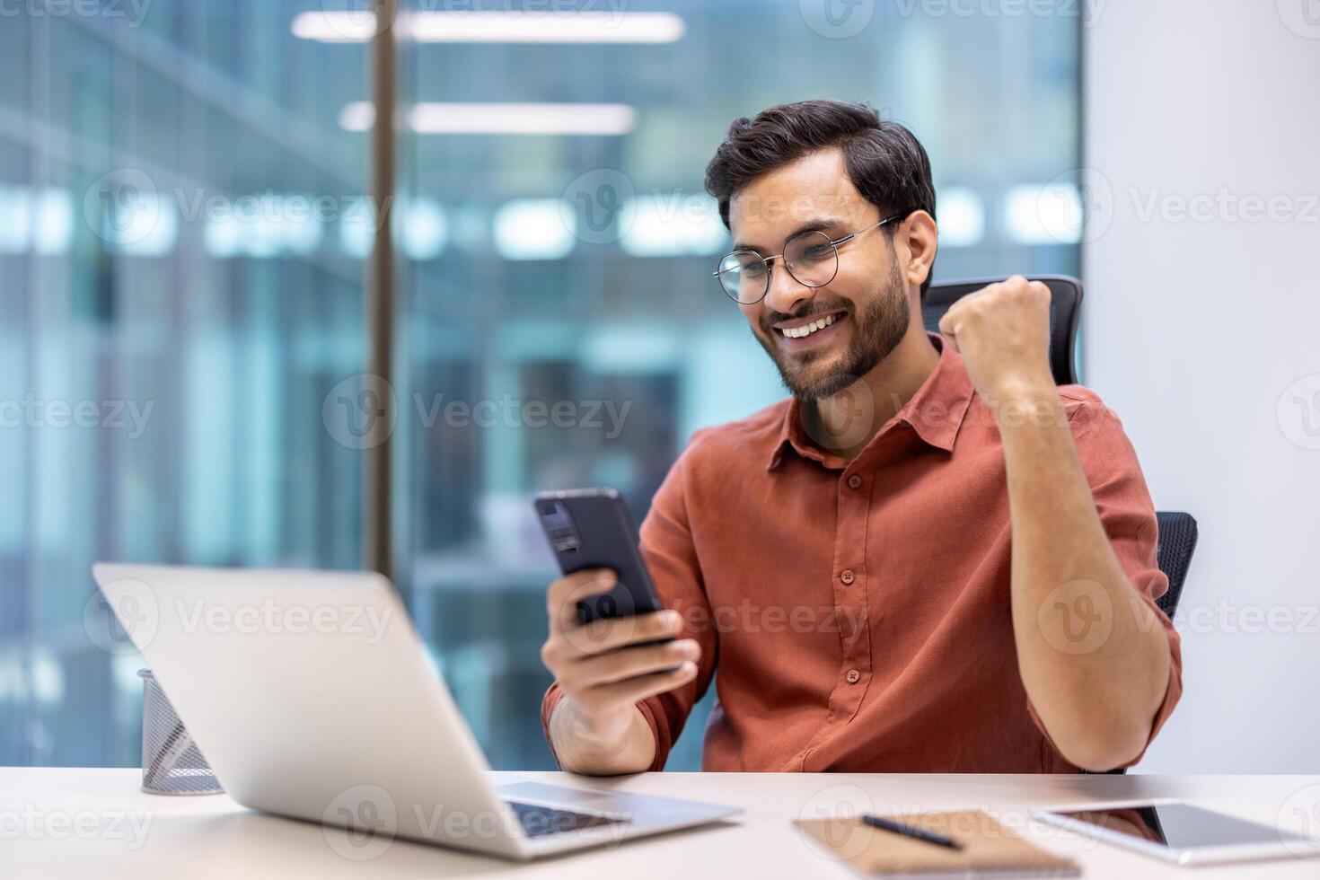Excited businessman celebrating success at work using smartphone. Young professional sitting at office desk with laptop, phone, and notebook, showing joyful expression and happiness. photo