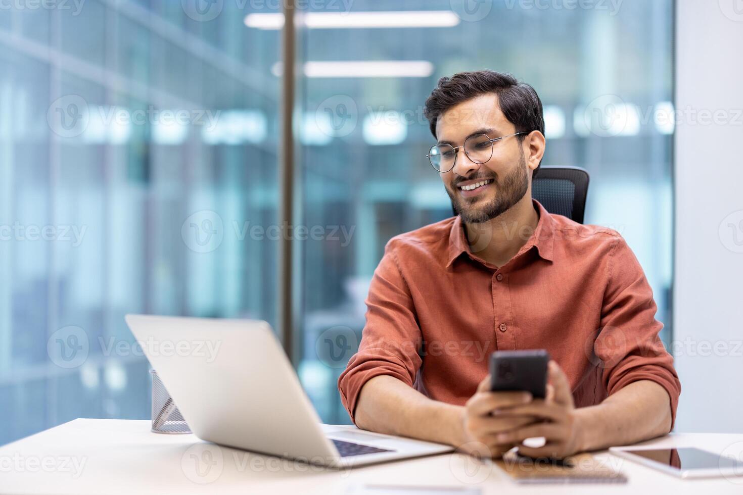 Businessman smiling while working on laptop and smartphone in modern office setting. Confident professional multitasking at bright workplace. Glass walls and contemporary design in background. photo