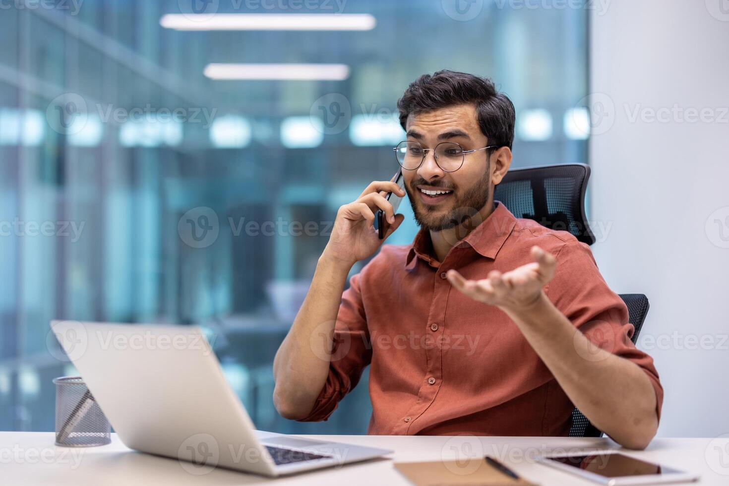 Confident man in office setting engaged in phone call while using laptop. Displays elements of technology, communication, business professionalism. Captures essence of modern work environment photo