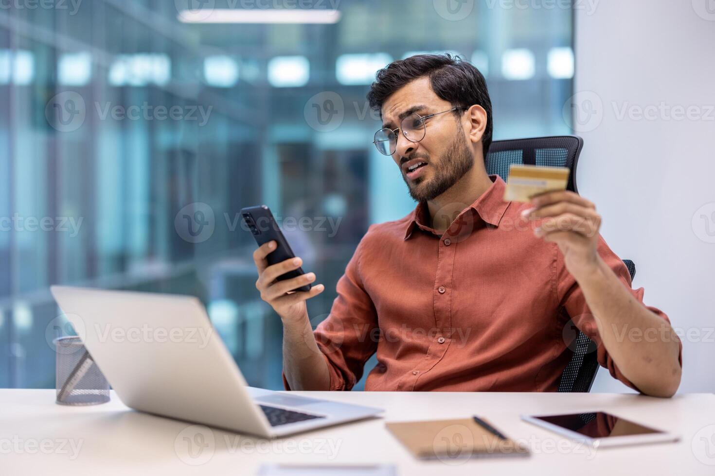 Businessman in modern office holding credit card, looking concerned about phone fraud. Laptop, notebook, and tablet on desk. Concept of online fraud, cyber security, and technology issues photo