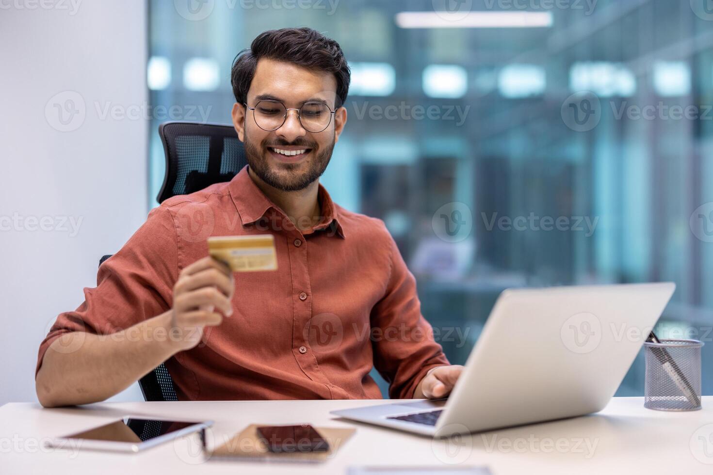 Smiling man using laptop for online shopping holding credit card. Modern office setting with phone and tablet on desk. Concept of e-commerce, technology, and finance. photo