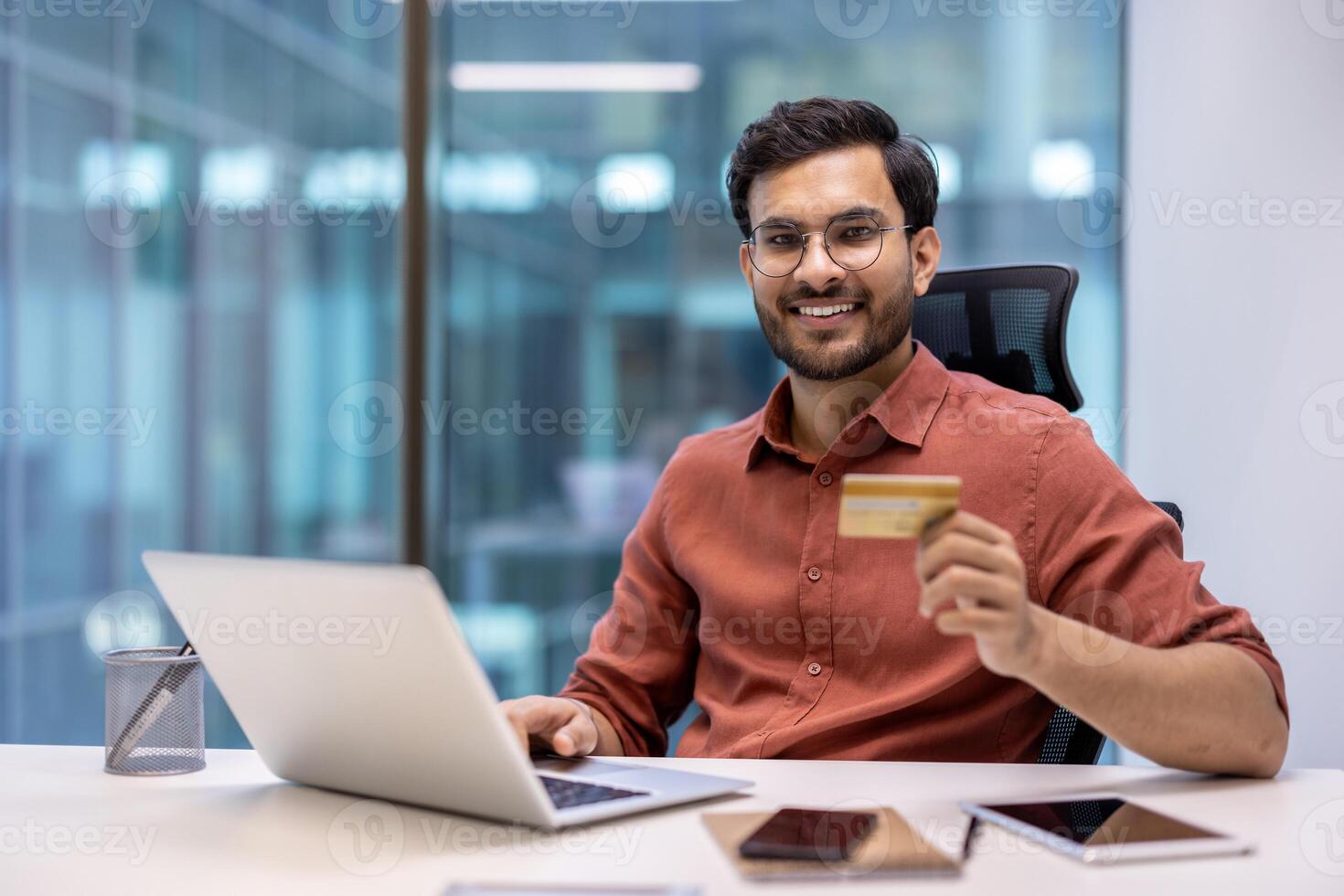 Business professional sitting at desk holding credit card using laptop in modern office. Engaged in online shopping or banking. Happy and confident expression. photo