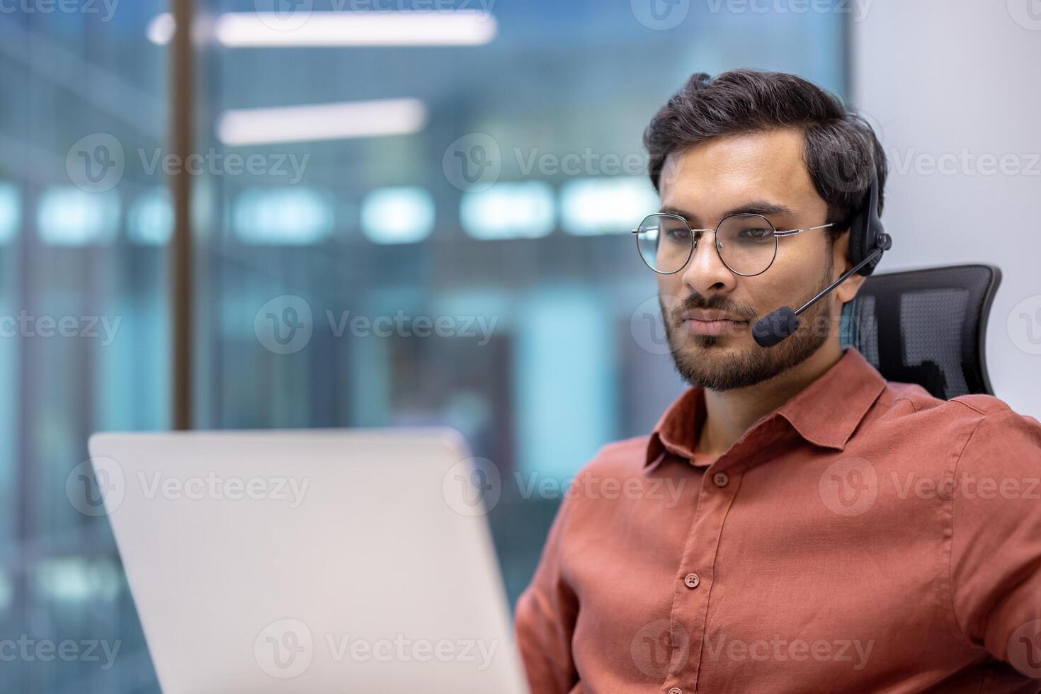 Young professional wearing headset working on laptop in modern office setting. Focused and engaged in online communication, displaying confidence. Formal attire suggests business environment. photo