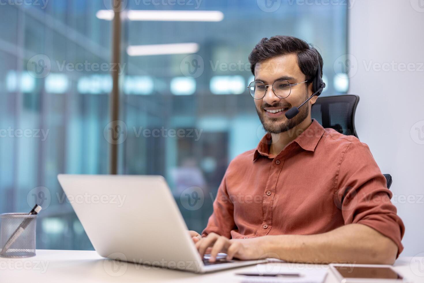 Smiling professional wearing headset working on laptop in modern office. Engaged in online communication, exemplifying technology's role in business. Reflects technology, and customer service. photo