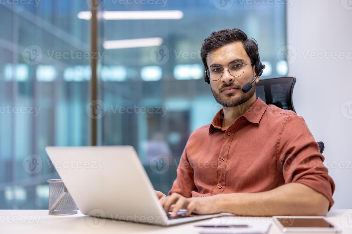 Latin American man businessman wearing glasses and headset working at office desk with laptop, exuding professionalism and focus. Multitasking skills emphasized, engaging in virtual communication. photo