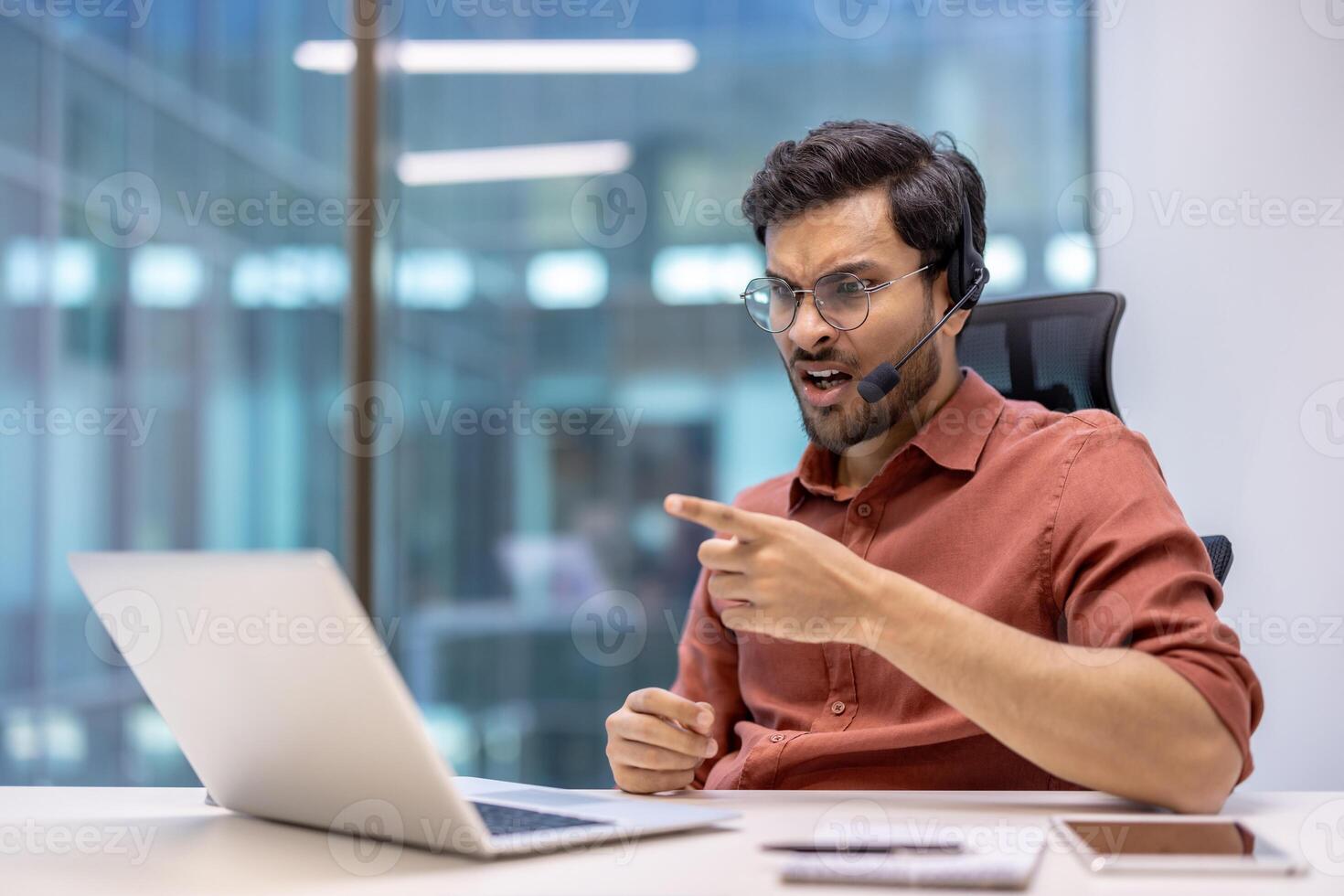 Latin American man businessman engaging in call using laptop and headset at office, expressing frustration. Technology, remote work, communication challenges, business setting depicted. photo