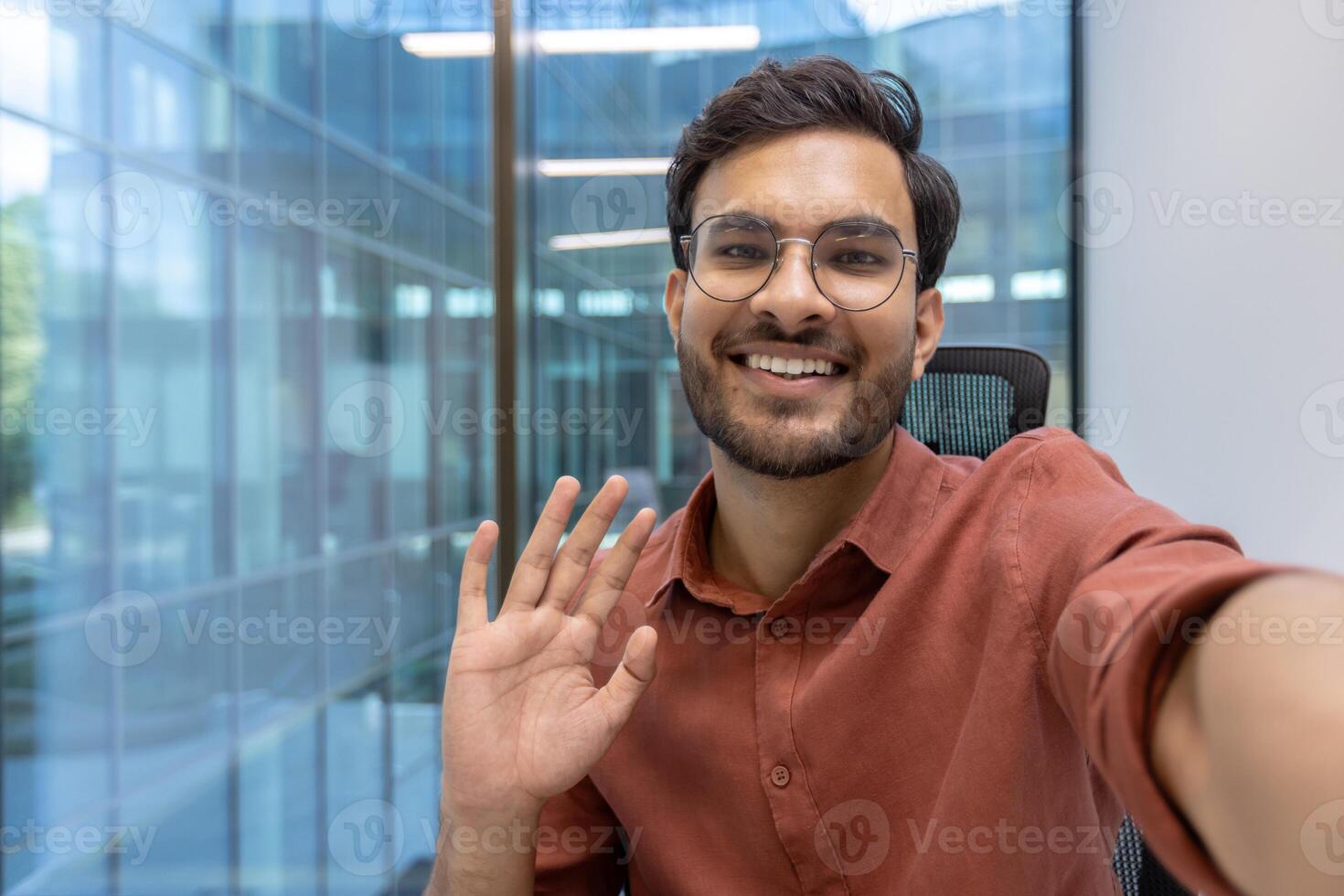Businessman wearing glasses smiling confidently during call in modern glass office. Man waving while using phone for virtual communication, representing connection, technology, entrepreneurship photo