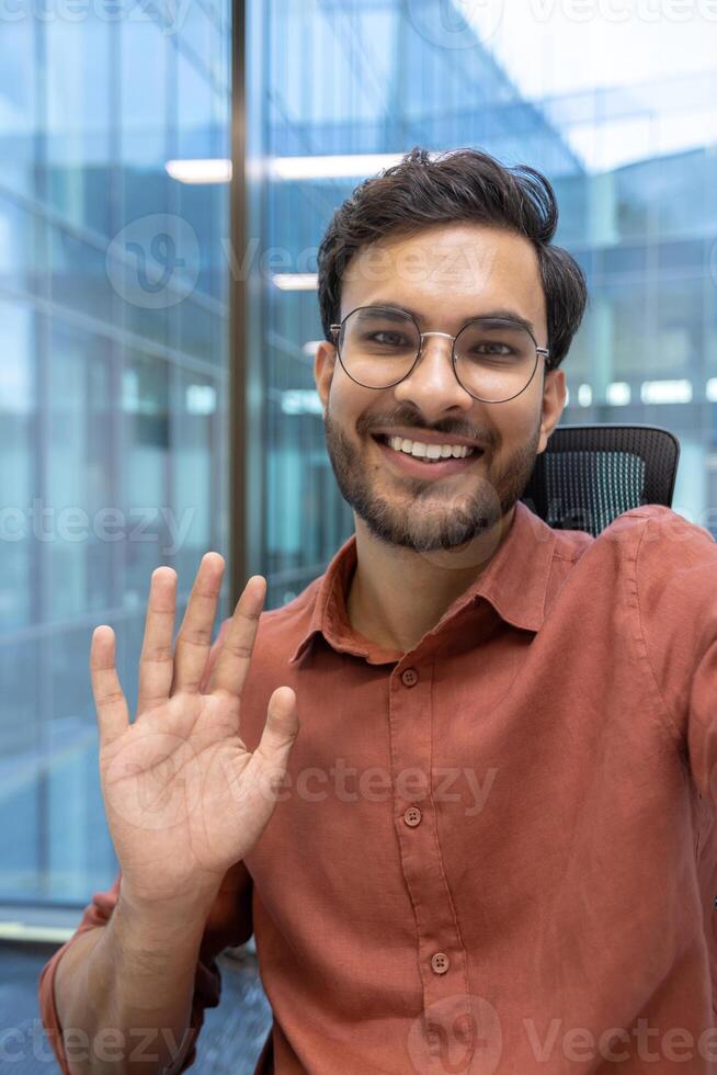 Cheerful young man with glasses waves during virtual meeting in modern office. Demonstrates concept of friendly communication, technology use, remote work, and professional interaction using phone in contemporary environment. photo
