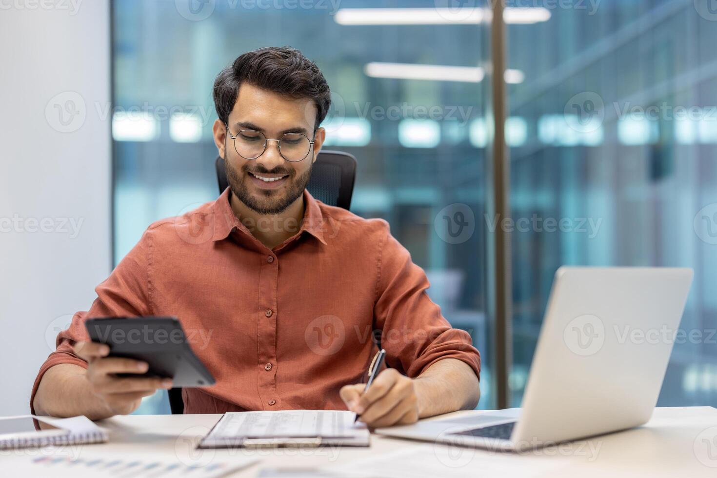 Smiling financier accountant reviews financial data with calculator and laptop. Professional efficiency displayed as he confidently tackles calculations and reports in modern office setting. Man exudes confidence and focus. photo