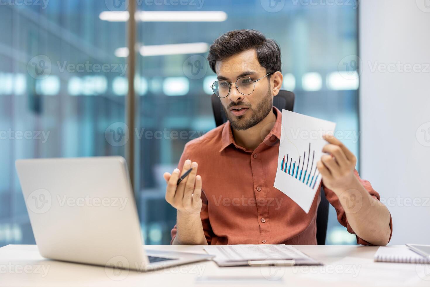 Businessman in office holding paper chart, explaining financial data during call on laptop. Focus on analysis, communication, technology. Reflects professional environment and office work photo