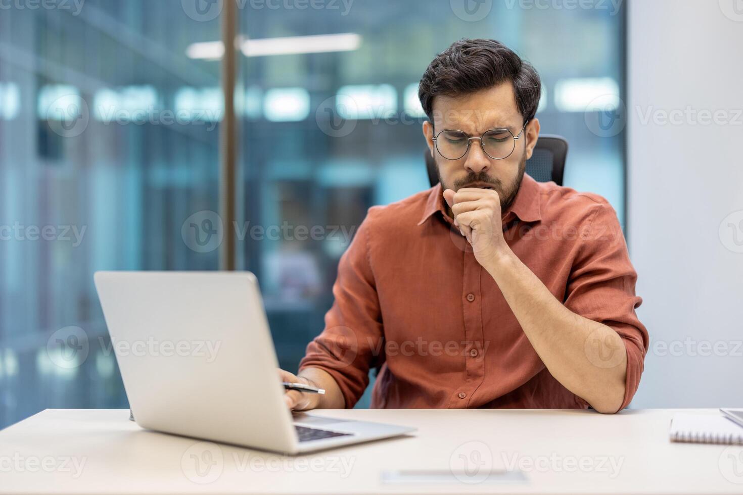 Male office worker sits at desk wearing glasses looking concerned, experiencing cough symptoms while using laptop. Workplace setting suggests professional environment photo