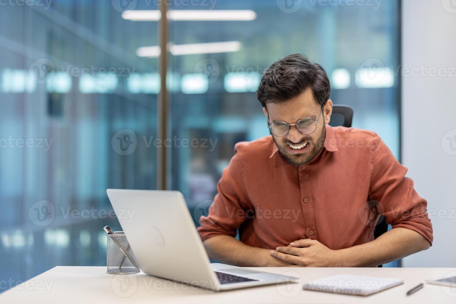 Business professional in office showing signs of abdominal pain and nausea while using laptop. Man wearing glasses, appearing stressed and uncomfortable, holding stomach. photo