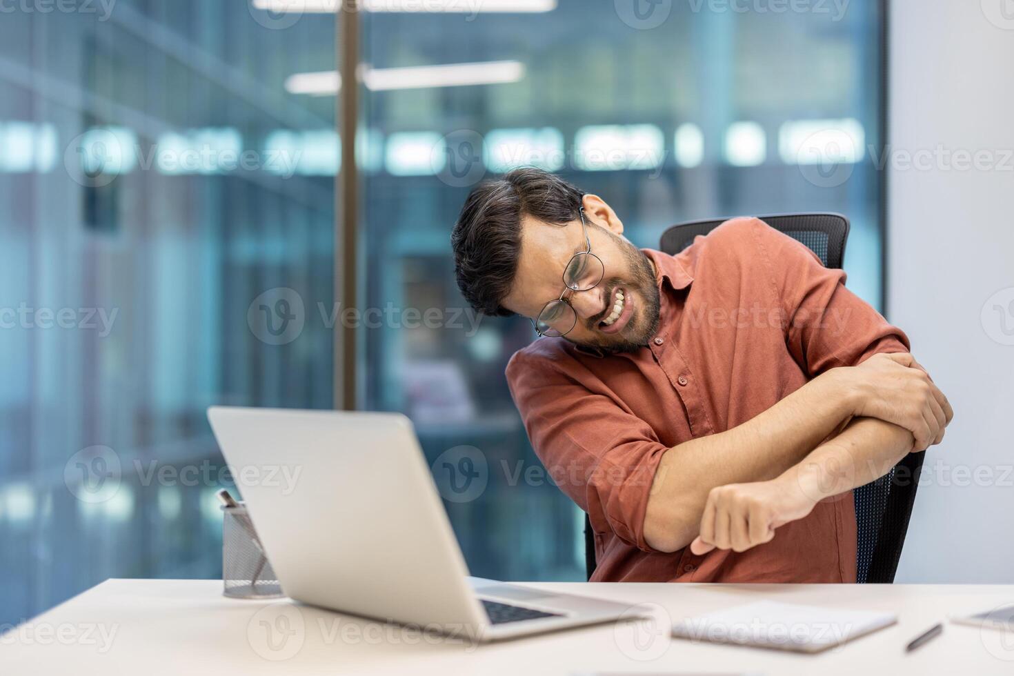 Man in office showing discomfort, experiencing arm pain while seated at desk using laptop. Expression of strain, reflecting common workplace health issues. Professional attire, modern workspace photo