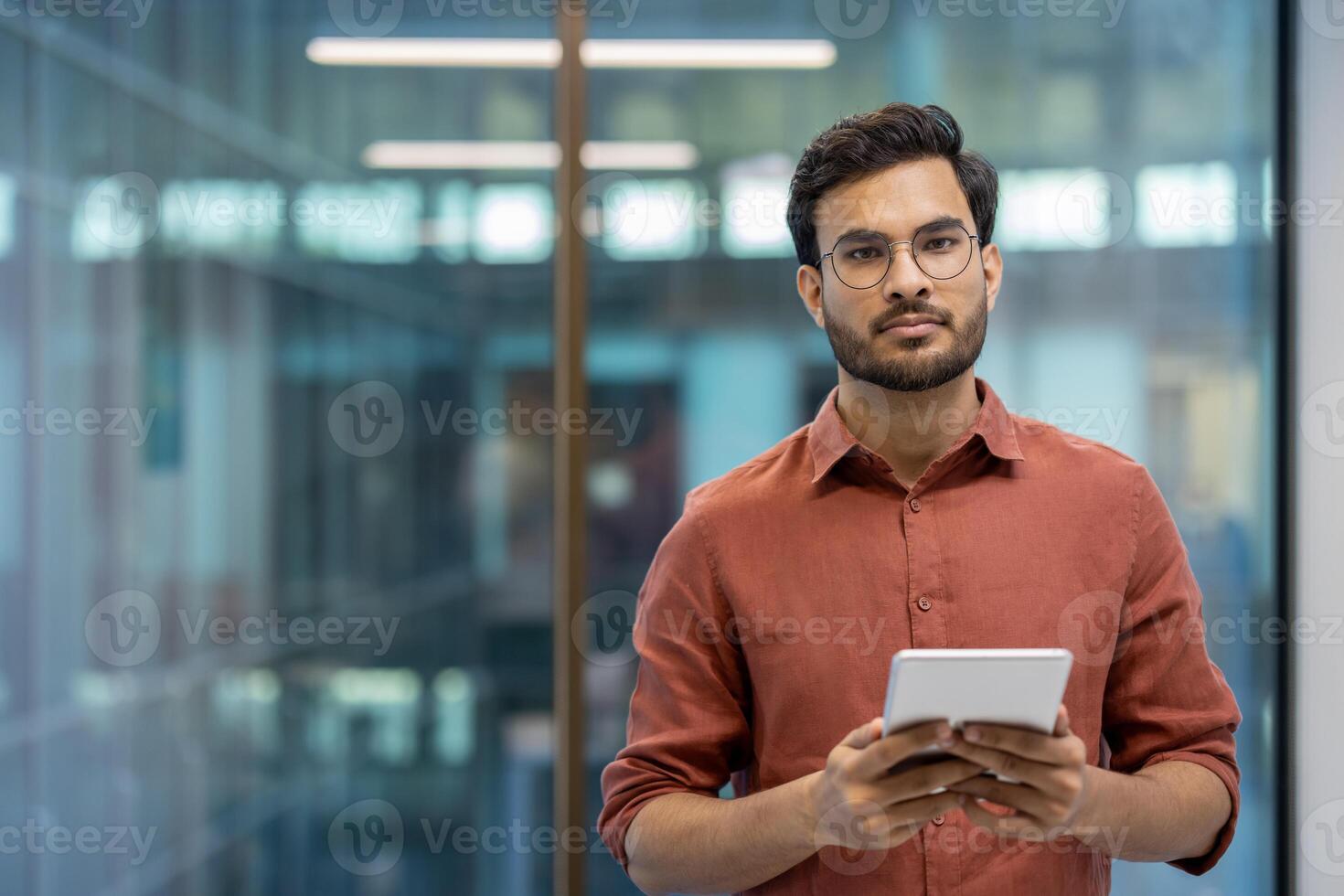 Indian programmer in modern office environment holding tablet device. Expression of confidence and professionalism. Suitable for concepts of technology, software development, and digital innovation. photo