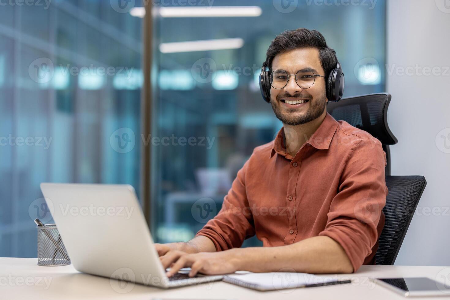 Young professional with glasses and headphones using laptop in modern office setting. Person smiling confidently while engaged in digital work. Office environment encourages creativity and focus photo