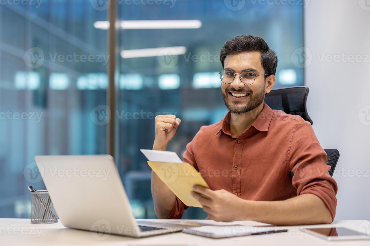 Happy businessman in office setting, seated at desk with laptop and documents, celebrating success. Bright, modern workplace conveys motivation, goal achievement, and positive working environment. photo