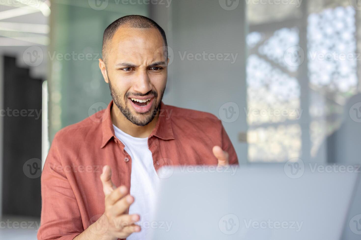 A young man looks surprised at his laptop during a call in a modern, bright room. He is reacting to something on the screen photo