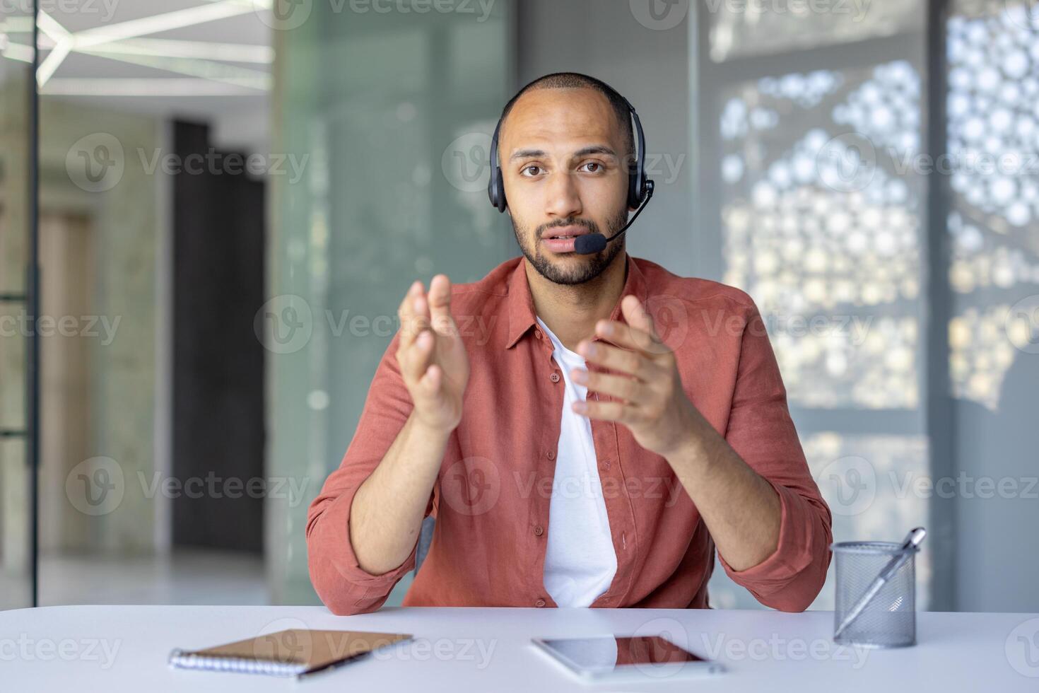 Young man with a headset having a call and gesturing with his hands in an office environment at a white desk photo